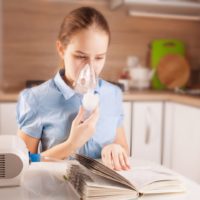 Girl with asthma using inhaler and reading book in the kitchen